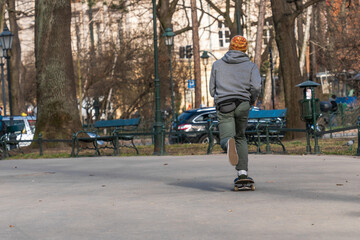 A young man vigorously rides a snowboard on an asphalt road through a town square in early spring. View from the back