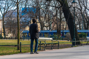 A young man in a short black coat holds a phone in his hand from which he listens to music in wireless headphones while walking in the city in early spring