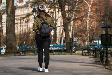 Close-up of a girl in a short jacket and sweatpants with a black bag on her back and a bottle of water walking along an asphalt road on a sunny day in early spring. Rear view horizontal orientation