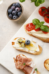Italian antipasto bruschetta - white bread slices with smoked ham, goat cheese, blueberries, mozzarella, tomatoes, basil on marble board, glass with lemon water