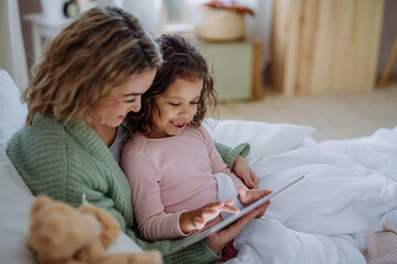 Happy mother with her little daughter lying on bed and using tablet at home.
