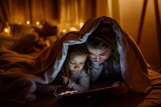 Happy Mother With Her Little Daughter Lying Under Blanket And Watching Movie On Tablet At Home.