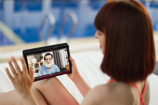 Vacations, Resort, Technology Concept. Over The Shoulder View Of Tablet Pc Screen. Back View Of Young Woman Relaxing At Pool And Having Video Call With Her Senior Bearded Grandfather, Waving Hand