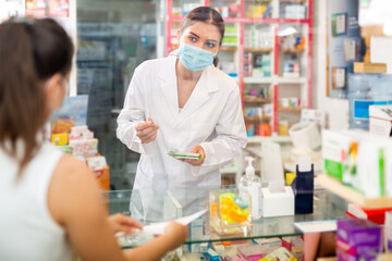Young female pharmacist in a protective mask, working in a pharmacy during the pandemic, takes an order for a medicine from .a buyer