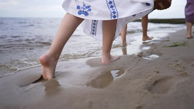 Barefoot feet of slim young women in traditional Ukrainian embroidered dresses walking in slow motion on sandy beach. Live camera follows unrecognizable ladies strolling outdoors