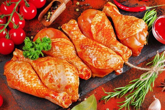 Barbecue marinated chicken dramstik on a black cooking board, with fresh rosemary leaves and fresh cherry tomatoes and various spices, close-up.Semi-finished product, Marinated product, quick cooking.