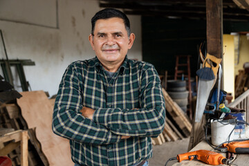 Fototapeta na wymiar Hispanic carpenter smiling proud in wood shop of him - Proud owner of carpentry shop standing with arms crossed