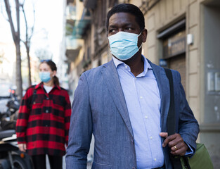 Fototapeta na wymiar Focused african american adult man wearing medical mask walking to work along city street in spring morning. New life reality during coronavirus pandemic