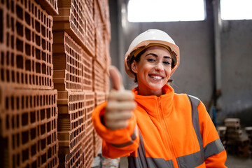 Female factory worker in safety equipment holding thumbs up for successful production of clay bricks for building industry.