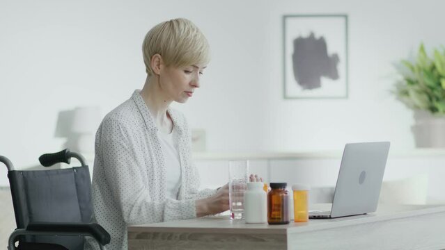 Mature woman with disability taking pills and drinking water, sitting in wheelchair and reading appointment on laptop