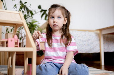 Montessori material. Child girl in pink T-shirt arranges wooden furniture in a doll house.