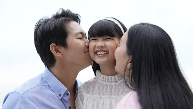 Holiday concept of 4k Resolution. Father and mother kissing daughter's cheek on the beach. Young Asian girl smiling happily. parents-child relationship.
