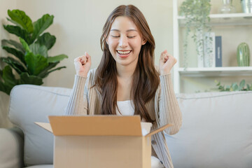 Happy excited, asian young woman, girl customer sitting on sofa at home, opening and unpacking cardboard box carton parcel after buying ordering present, shopping online, delivery service concept.