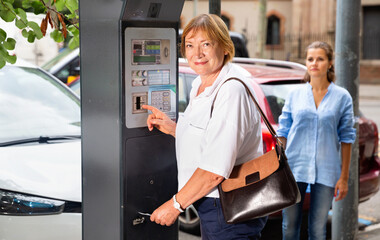 Cheerful positive middle aged woman using parking machine to pay for car parking on summer city street