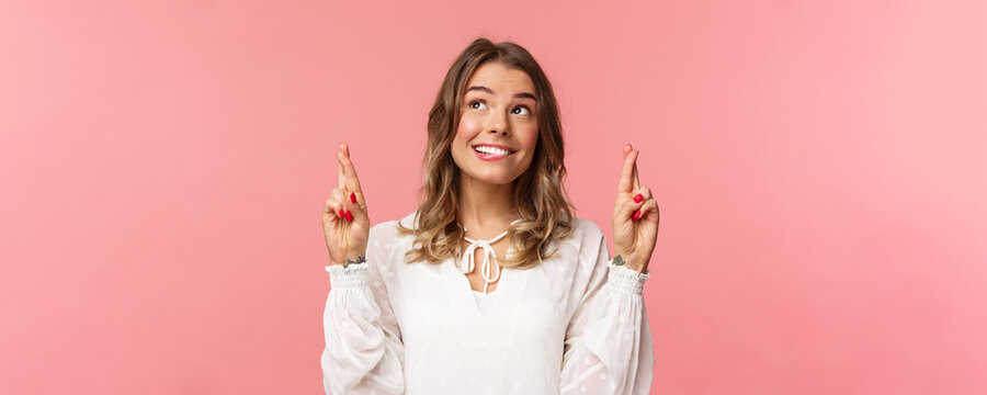Close-up portrait of hopeful, optimistic attractive woman in white dress, bite lip and smiling as daydreaming, hope dream come true, making wish or praying, standing pink background