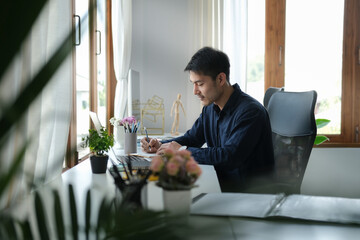 Young asian businessman sitting in bright modern workplace and working with laptop computer.
