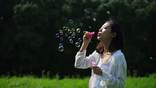 Charming Chinese girl standing outdoors, blowing soap bubbles smiling