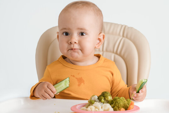 A baby on a feeding chair is eating a cucumber. The concept of proper baby food.