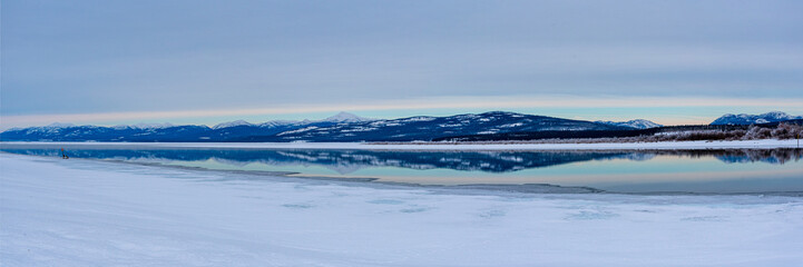 Panoramic winter views with mountain reflection in calm water. Snow capped peaks in background. 