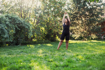 Over-weighted female holding hands up stretching doing yoga enjoying the moment on green grass on backyard of cottage with wooden house and trees in background. Body positivity. Equality. 