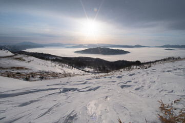 雪に覆われた斜面の向こうの靄に覆われた湖と曇り空に輝く太陽。日本の北海道の美幌峠の風景。