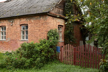 Fototapeta na wymiar Picture of rural lovely sweet old red-brick house with grey tile roof, big windows, small cute fence and blue mailbox, green plants growing on facade of dwelling and in front of it