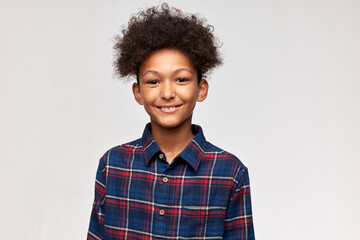 Head and shoulders studio shot of sweet handsome dark-skinned male kid of 13 with cute lovely toothy smile, dressed in stylish clothes, wearing fluffy afro haircut, isolated on white wall © Anatoliy Karlyuk