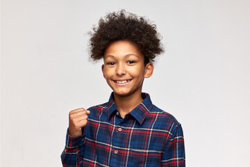 Studio shot of happy enthusiastic dark-skinned boy in flannel shirt showing yes sign with clenched fist, smiling isolated on white wall. Body language, gestures, emotions and feelings