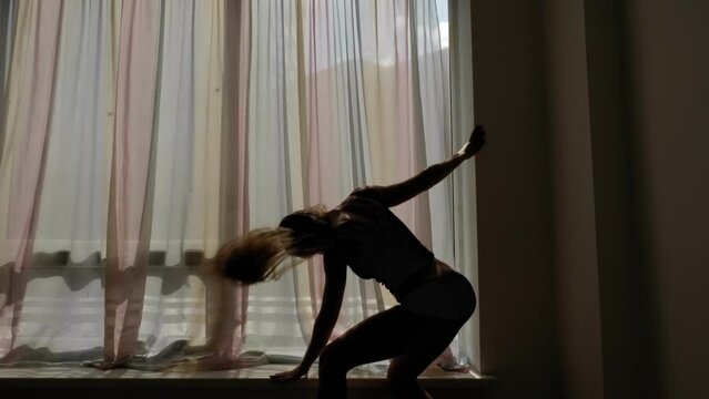 The silhouette of a sexy woman in underwear and striptease shoes who dances a sensual dance near a large window with long curtains.