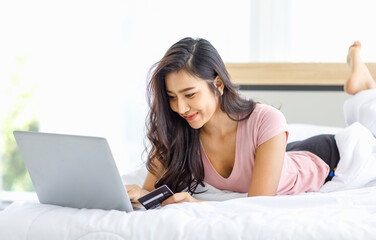Asian young happy cheerful beautiful female model in casual pajama outfit lying down smiling hold credit card on bed in bedroom browsing surfing internet shopping online via laptop notebook computer