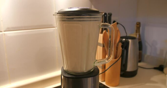 View of homemade oat and milk mix grinding in a blender making oat milk in the kitchen with white tile in the background. Light falling on the blender.