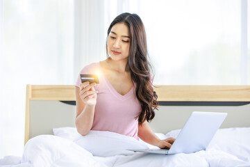 Asian young pretty female shopper in casual pajama waking up sitting smiling under blanket on bed in bedroom holding looking at credit card with light flare effect shopping online via laptop computer