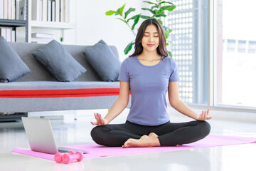 Asian young happy peaceful calm female model in casual sporty outfit sitting crossed legs in lotus position on yoga mat learning studying online meditation class via laptop computer in living room - obrazy, fototapety, plakaty