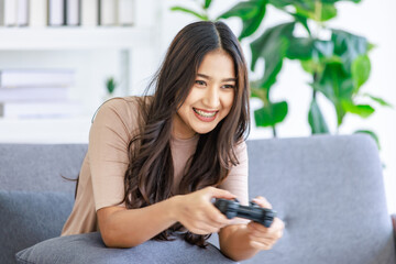 Asian young beautiful cheerful happy exciting female gamer model sitting on cozy couch smiling holding joystick controller playing fun game via console gaming platform online in living room at home