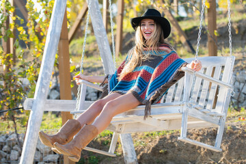 Fashion woman in boho style on wooden bench. Beauty blonde hipster girl in hat relaxing outdoors on porch swing in the nature. Outdoor portrait of a smiling happy girl, cheerful girl laughing at park.