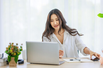 Asian young beautiful happy professional successful businesswoman designer sitting smiling at workstation desk using laptop notebook computer and smartphone working remotely online at home office