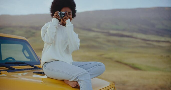 Beautiful young black african american woman relaxing and taking pictures at sunset with vintage camera on epic road trip, adventure travel lifestyle