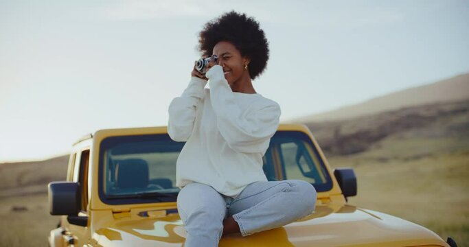 Beautiful young black african american woman relaxing and taking pictures at sunset with vintage camera on epic road trip, adventure travel lifestyle