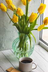 Morning coffee and a bouquet of yellow tulips in a glass vase on a light background by the window