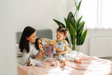 Mom and little daughter are sitting on the bed and playing with dolls. Motherhood, caring, time...
