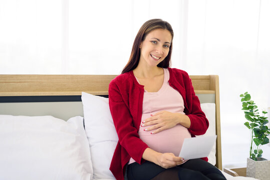 Pregnant woman sitting on bed and looking at her ultrasound image of her child in her belly.