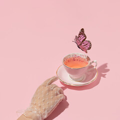 Woman hand reaching vintage cup of tea and butterfly flying above on pastel pink background. 80s, 90s retro aesthetic spring or summer concept. Minimal fashion romantic idea.