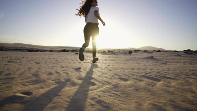 Young black woman running with sandals in sand dunes desert the sunset by scenic wavy texture sand surface. Close up woman legs with sunshine on background. Inspirational travel adventure slow motion
