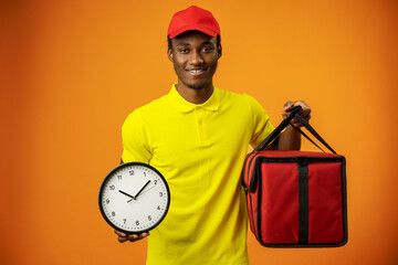 Smiling african american male courier holding clock and box on orange background