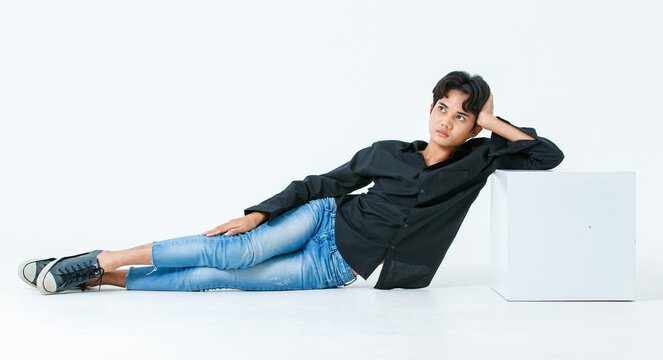 Portrait isolated cutout studio shot of Asian young LGBTQ gay glamour male model in casual black shirt and jeans outfit sitting stretching posing gesturing on floor look at camera on white background