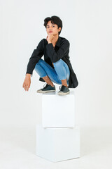 Portrait studio shot of Asian young LGBT gay bisexual homosexual topless male fashionable model in casual black shirt and denim jeans sitting on square boxes posing look at camera on white background