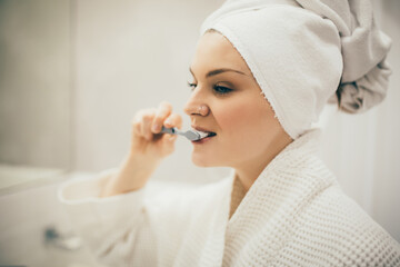 Young woman wearing white bathrobe and tower on head brushing her teeth using toothbrush and paste.