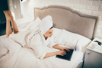 Young woman freelancer working from home using her laptop, lying in bed in white bathrobe and towel on head.