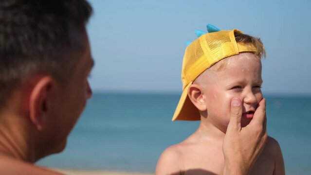 A father applies protective cream to his son's face at the beach. A man's hand applies sunscreen lotion on a child's face. Cute little boy with sunscreen by the sea. Copy space.