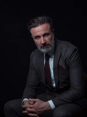 Portrait of adult businessman wearing trendy suit and sitting in modern studio on stylish chair against the black background